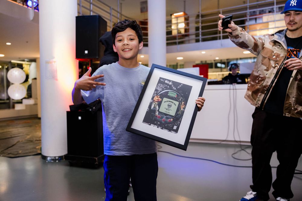 Een jongetje poseert met een lijst in zijn handen, waarin een poster zit. Rechts van hem staat een jonge man die in de lucht wijst, met een dj op de achtergrond.