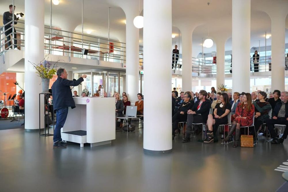 Spreker houdt toespraak achter een wit katheder voor een aandachtig publiek in het Glaspaleis van SCHUNCK, omringd door hoge witte zuilen en moderne architectuur.