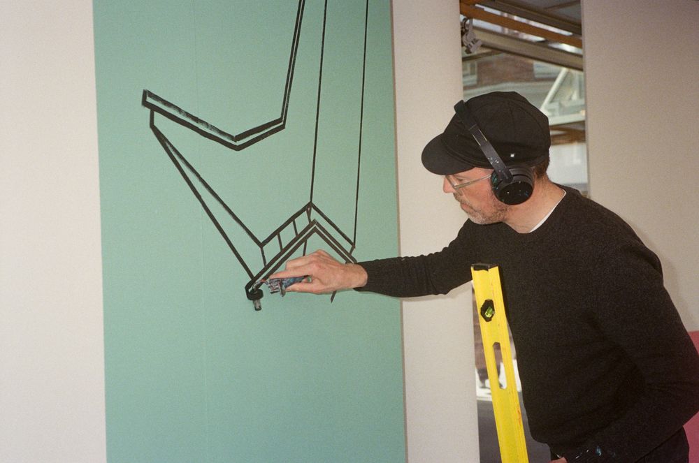 Kunstenaar Jeroen Erosie brengt met een marker een geometrische tekening aan op een lichtgroene wand tijdens de opbouw van een tentoonstelling.