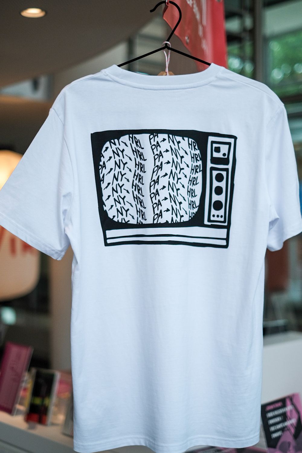 Een wit T-shirt met een zwart-witte illustratie van een televisie op de achterkant. Op het scherm staan herhalende teksten als “NY”, “HRL” en “PA” gedrukt in golvende lijnen. Het shirt hangt aan een zwarte kledinghanger in een moderne binnenruimte.