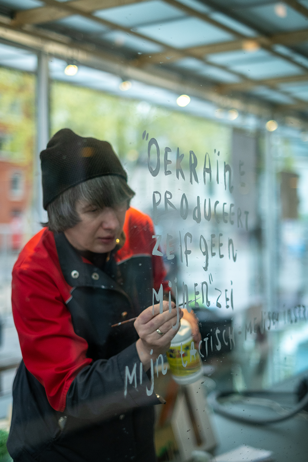 Kunstenaar in rood-zwarte jas schrijft met witte verf een tekst in het Nederlands op een glazen wand, binnen een lichte ruimte met grote ramen.