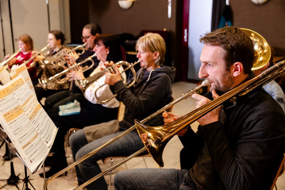 Volwassen muzikanten spelen samen op koperblaasinstrumenten tijdens een orkestrepetitie. Op de voorgrond bespeelt een man een trombone, terwijl de overige deelnemers geconcentreerd hun partij volgen vanaf de muziekstandaards.
