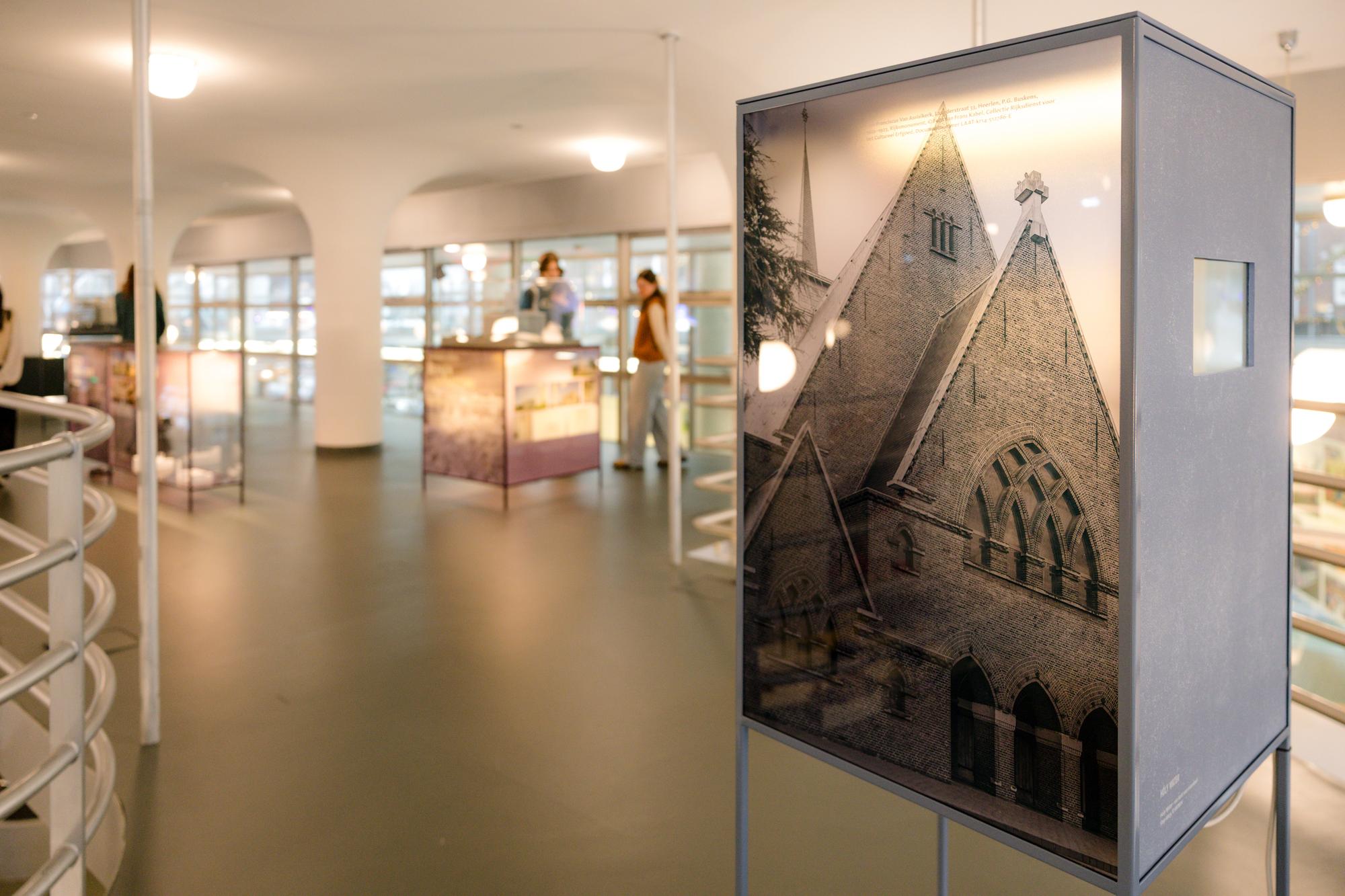 Tentoonstellingsruimte met een staand paneel waarop een zwart-witfoto van een kerkgevel te zien is, met vitrines en bezoekers op de achtergrond.