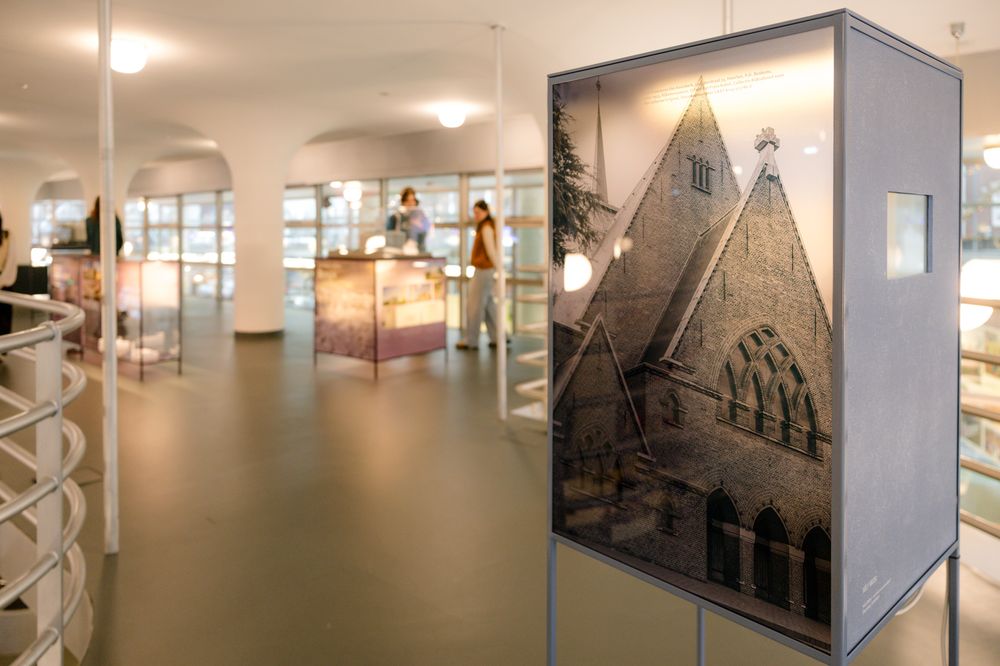 Tentoonstellingsruimte met een staand paneel waarop een zwart-witfoto van een kerkgevel te zien is, met vitrines en bezoekers op de achtergrond.