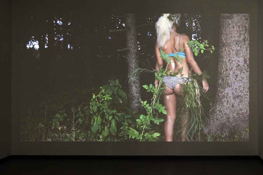 Videoprojectie waarin een vrouw in een bikini door het bos loopt.
