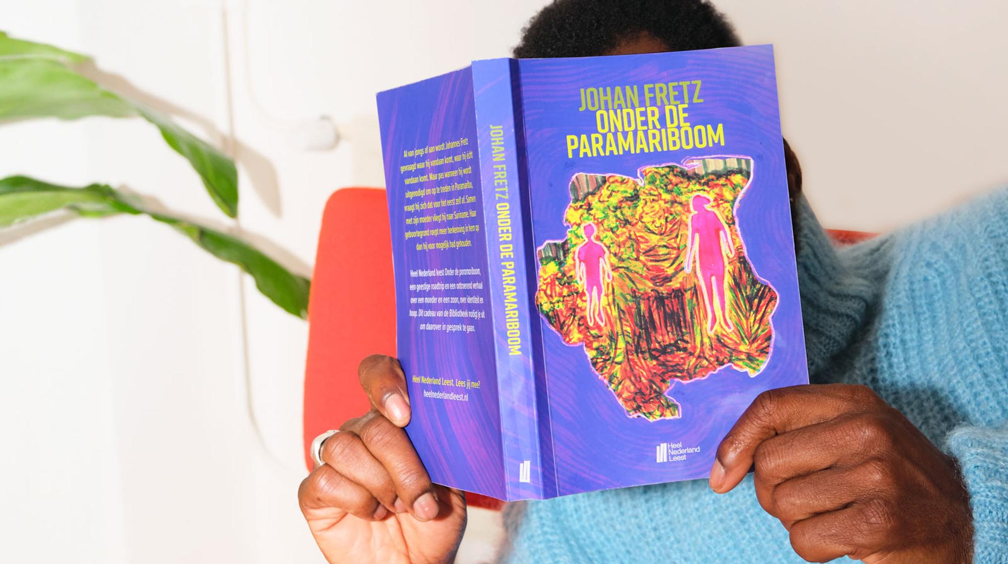 Persoon leest het boek 'Johan Fretz - Onder de Paramariboom', met een kleurrijke, abstracte omslag waarop twee menselijke figuren staan.