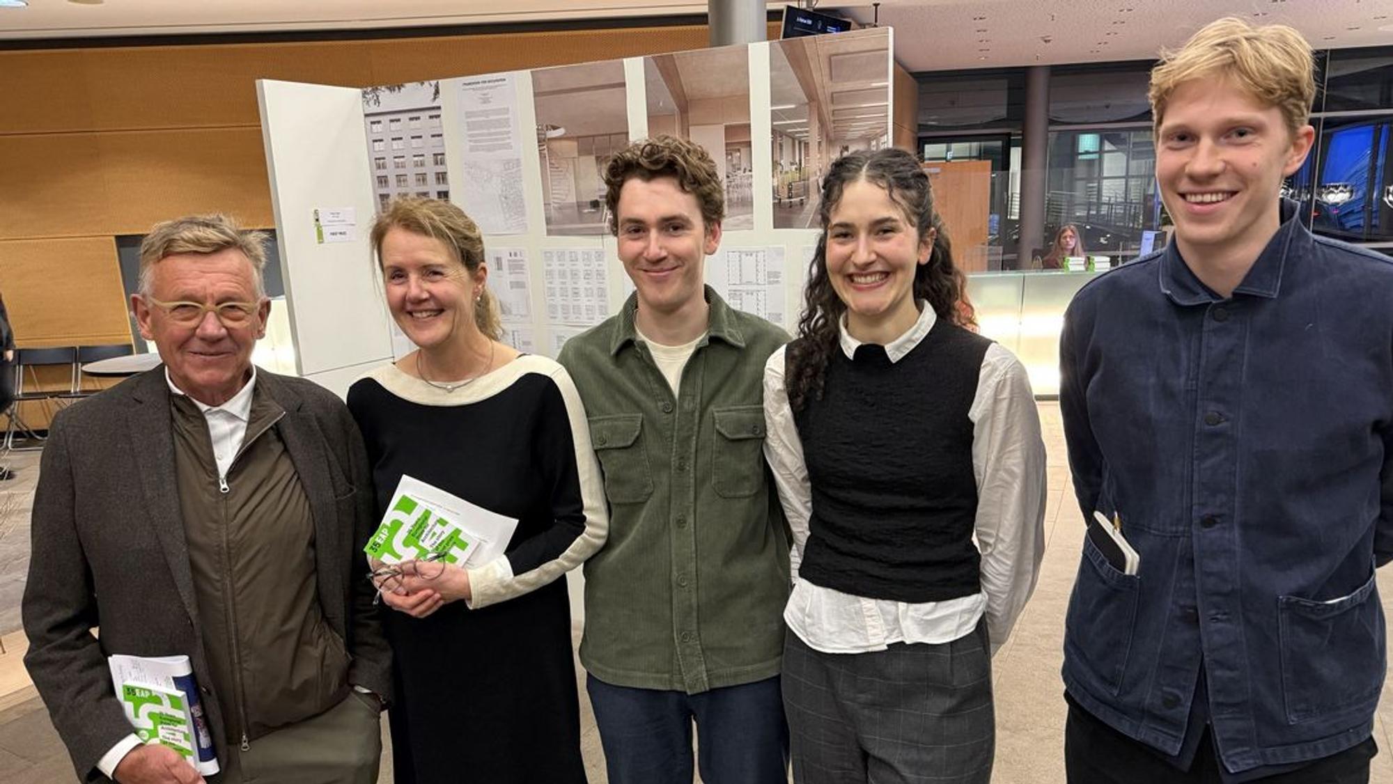 Groep van vijf mensen staat naast elkaar en glimlacht in een tentoonstellingsruimte met panelen op de achtergrond.