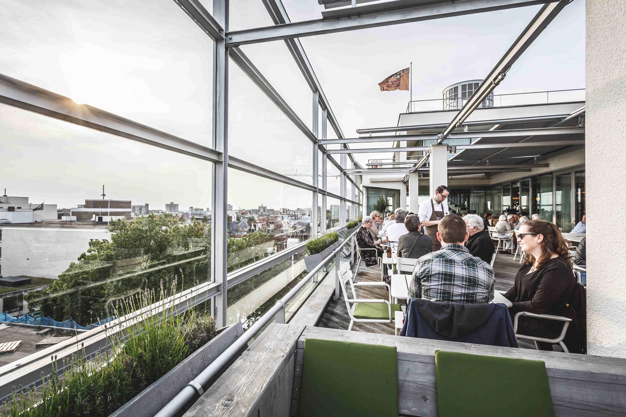 Dakterras van het SCHUNCK Glaspaleis, met tafels en stoelen van het restaurant en bezoekers. 