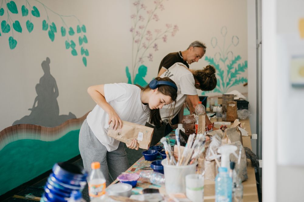 Drie kunstenaars werken geconcentreerd aan een schilderproject bij SCHUNCK in Heerlen, met schildermaterialen op tafel en muurschilderingen van planten en een silhouet van een zittende figuur op de achtergrond.