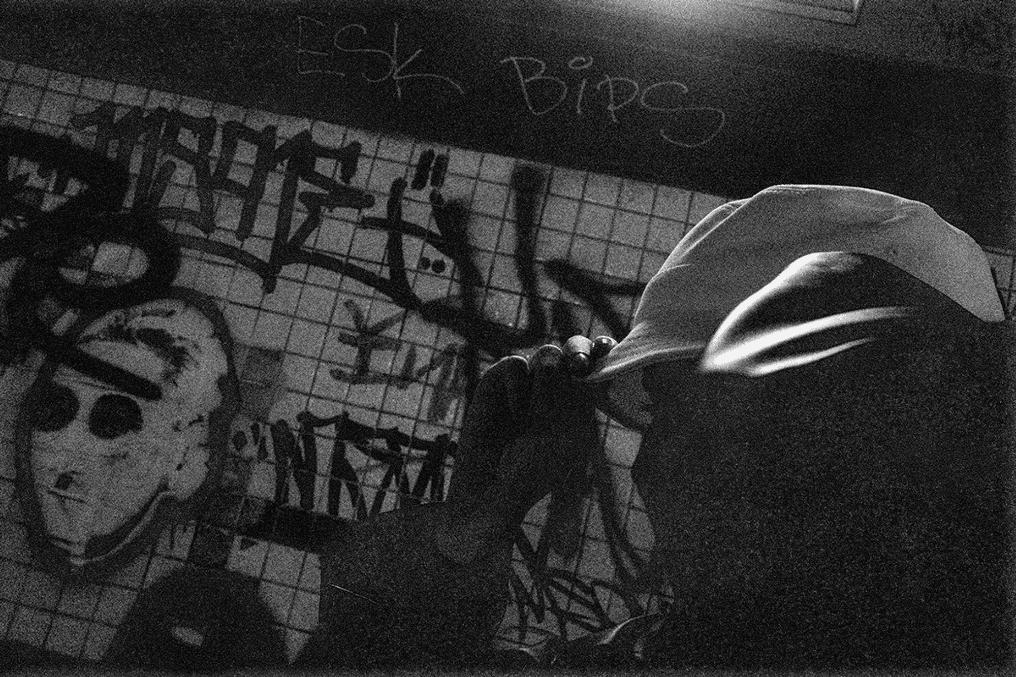 Zwart-witfoto van een persoon die een pet vasthoudt in een duistere omgeving, met op de achtergrond een muur vol graffiti en een getekend masker.
