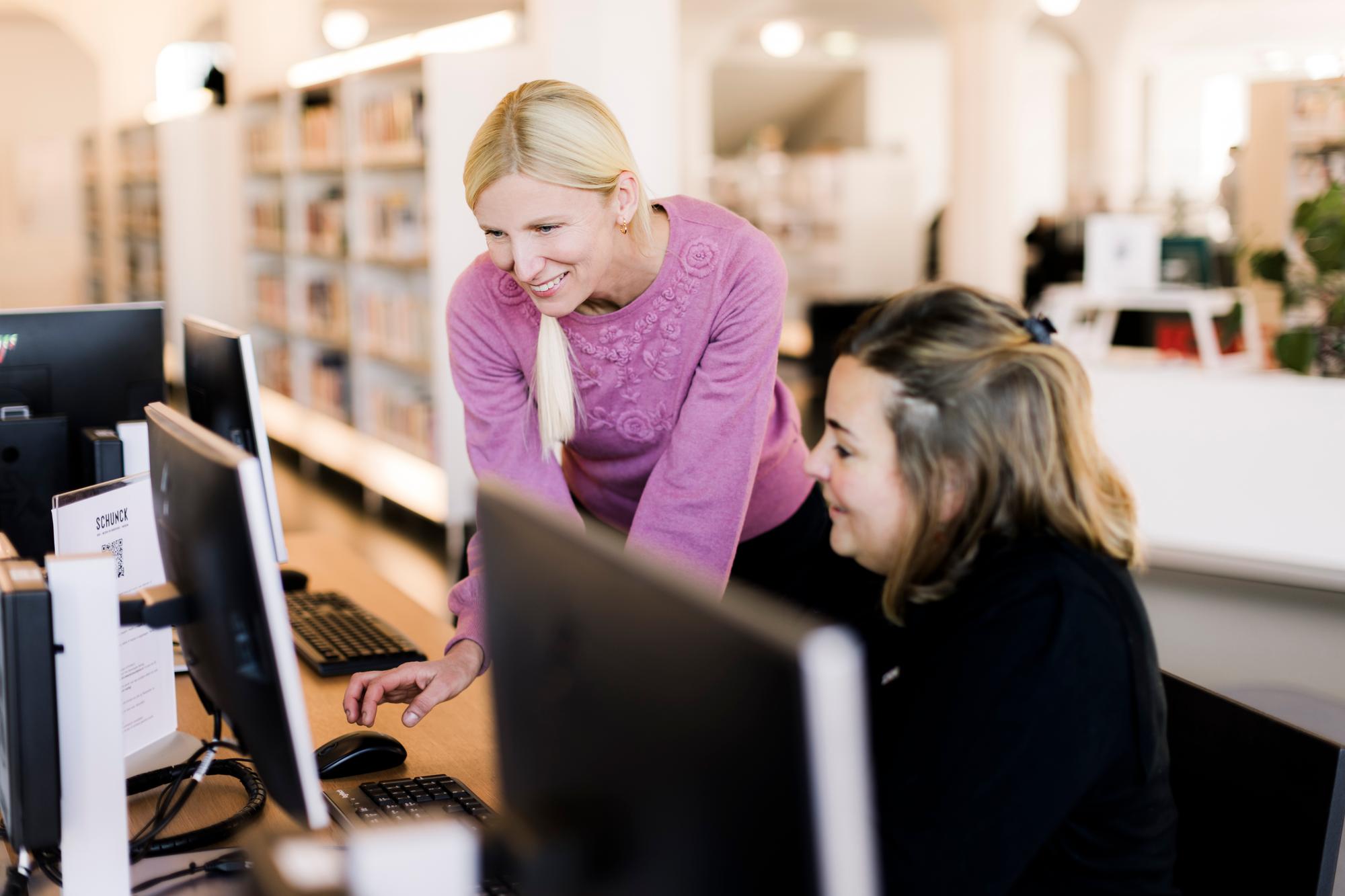 Twee personen werken samen achter een computer in een bibliotheekomgeving. Op de achtergrond zijn boekenrekken zichtbaar en meerdere computerschermen staan op de werkplek.