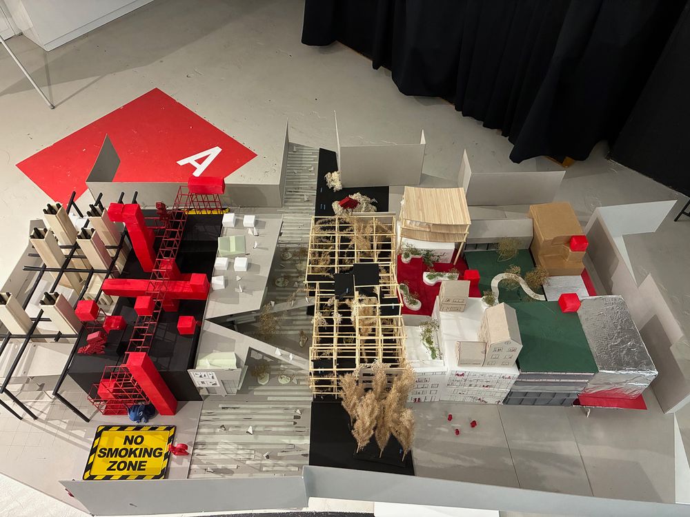 Bovenaanzicht van een grote architectuurmaquette met verschillende ontwerpen voor een bouwblok in Heerlen. De maquette bestaat uit gebouwen, rode constructies, houten raamwerken, groene vlakken en kleine bomen.