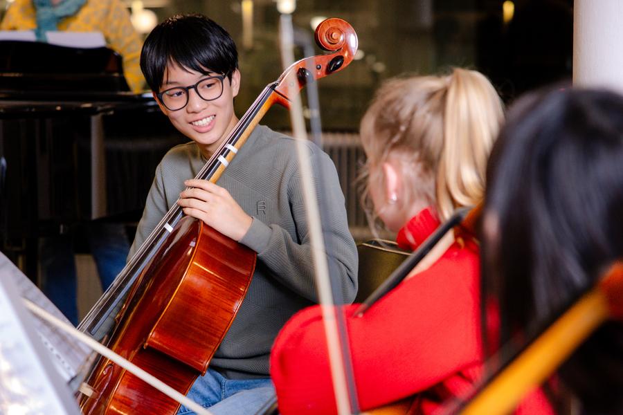 Een groep jonge muzikanten zit in een kring met cello’s tijdens een repetitie of muziekles. Op de voorgrond bespelen meerdere deelnemers hun instrument, terwijl op de achtergrond een pianist zichtbaar is achter een vleugel.
