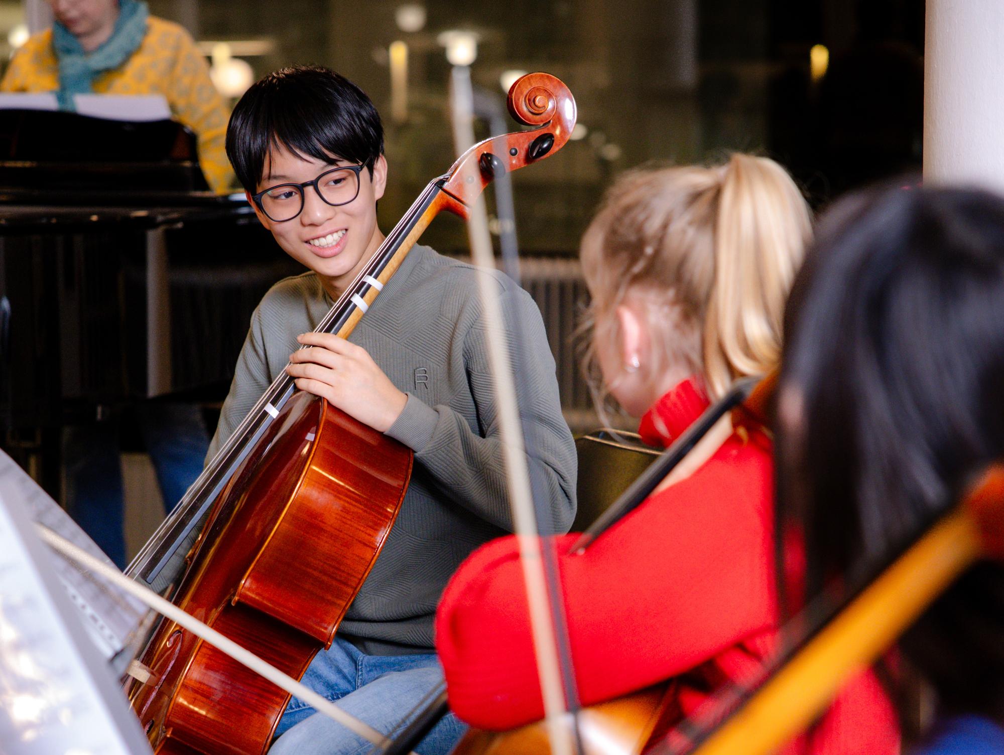 Een groep jonge muzikanten zit in een kring met cello’s tijdens een repetitie of muziekles. Op de voorgrond bespelen meerdere deelnemers hun instrument, terwijl op de achtergrond een pianist zichtbaar is achter een vleugel.
