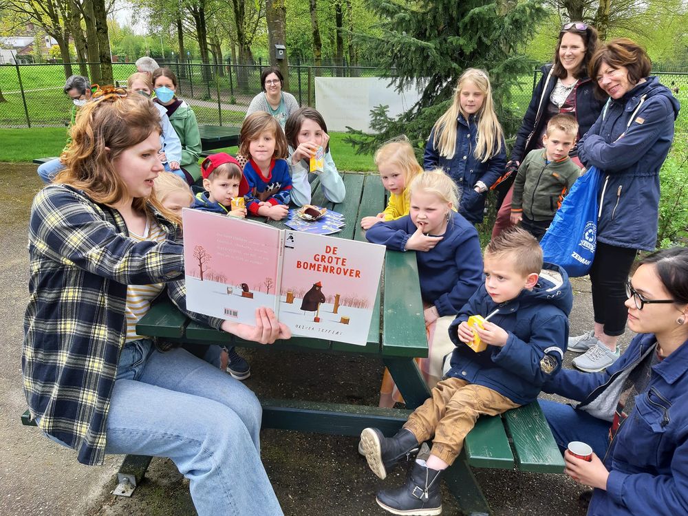 Groep kinderen en volwassenen luistert buiten naar een voorlezer met het boek 'DE GROTE BOMENROVER', zittend rond een groene picknicktafel met bomen en een hek op de achtergrond.
