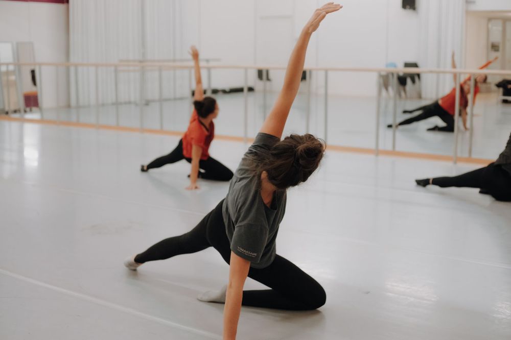 Drie dansers liggen op de vloer in de danszaal, met een been gestrekt en een arm in de lucht, terwijl hun andere arm hen ondersteunt.