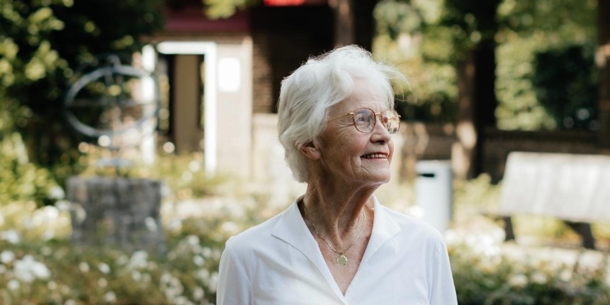 Foto van Irene Borghans, genomen in een tuin, met op de achtergrond een bankje, kunstwerk en planten. 