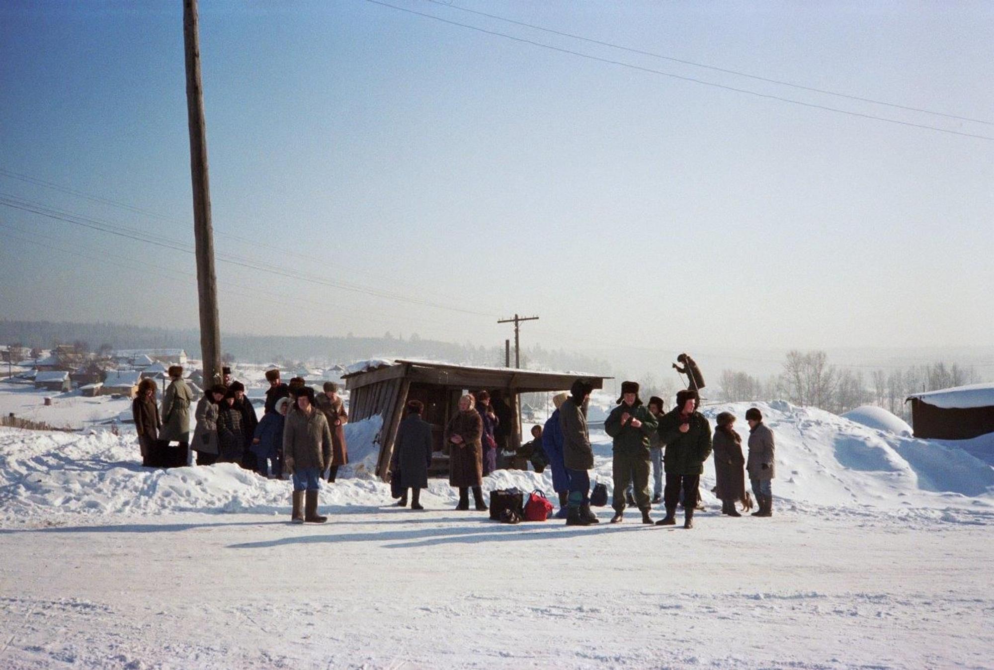 Een groep mensen wacht bij een bushalte in een besneeuwd Siberisch landschap. Ze dragen dikke winterjassen en bontmutsen tegen de kou.