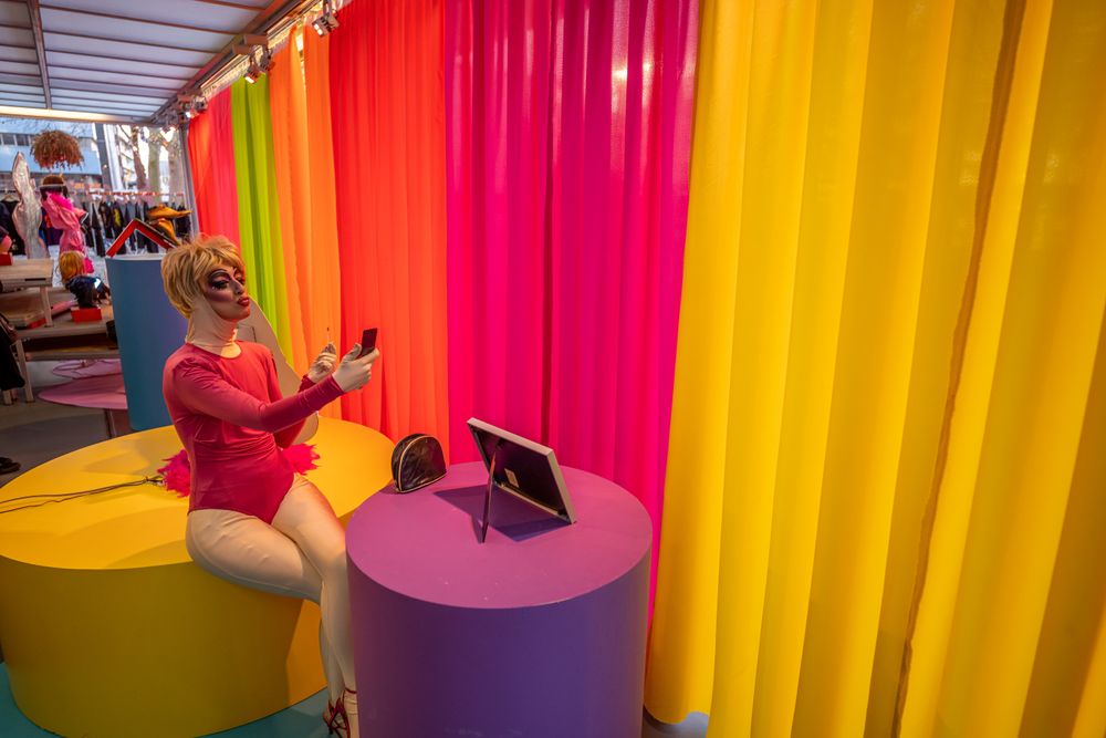 Dragqueen in roze outfit zit op een gekleurde ronde tafel en maakt een selfie, tegen een achtergrond van felgekleurde gordijnen in geel, roze, oranje en groen.