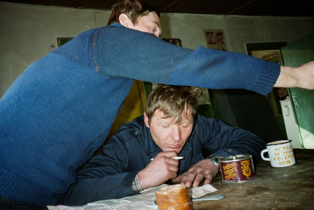 Fotografie 'At the Mines' van Bertien van Manen (1994), Apanas, Siberië. Twee mannen zitten aan een tafel; de linker man buigt over de rechter, die een pen in de hand houdt. Op de tafel staan mokken.