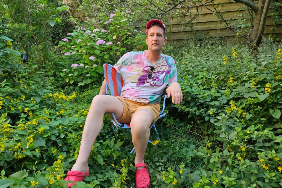 Maarten Bel, een man, zit ontspannen op een klapstoeltje in een weelderige tuin vol groene planten en gele bloemen. Hij draagt een kleurrijk T-shirt, beige korte broek, roze Crocs en een roze pet. Op de achtergrond zijn planten en een houten schutting te zien.