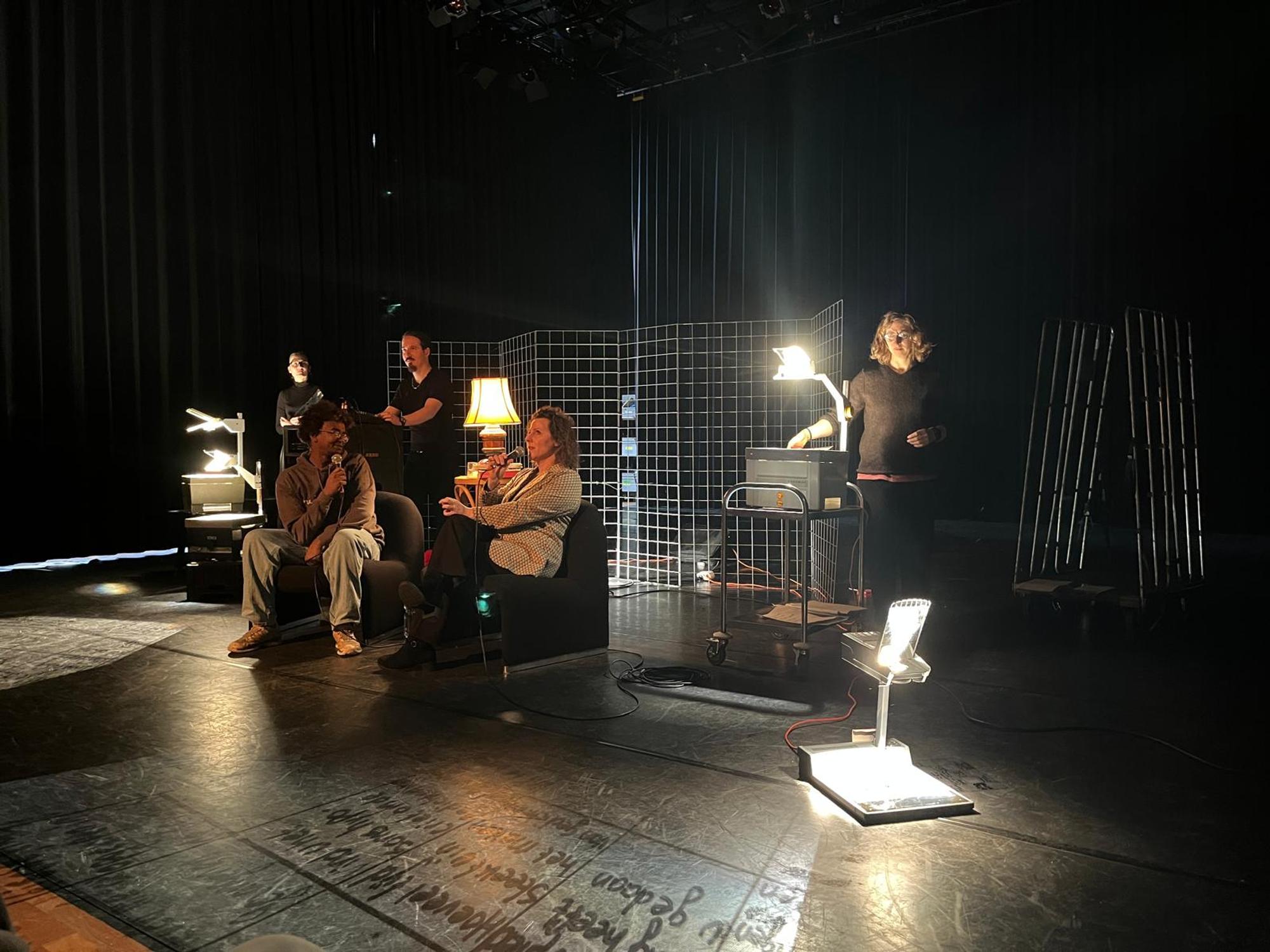 Repetitiesetting op een theaterpodium waar een dichter en gespreksleider in stoelen praten, omringd door lampen en projectoren.