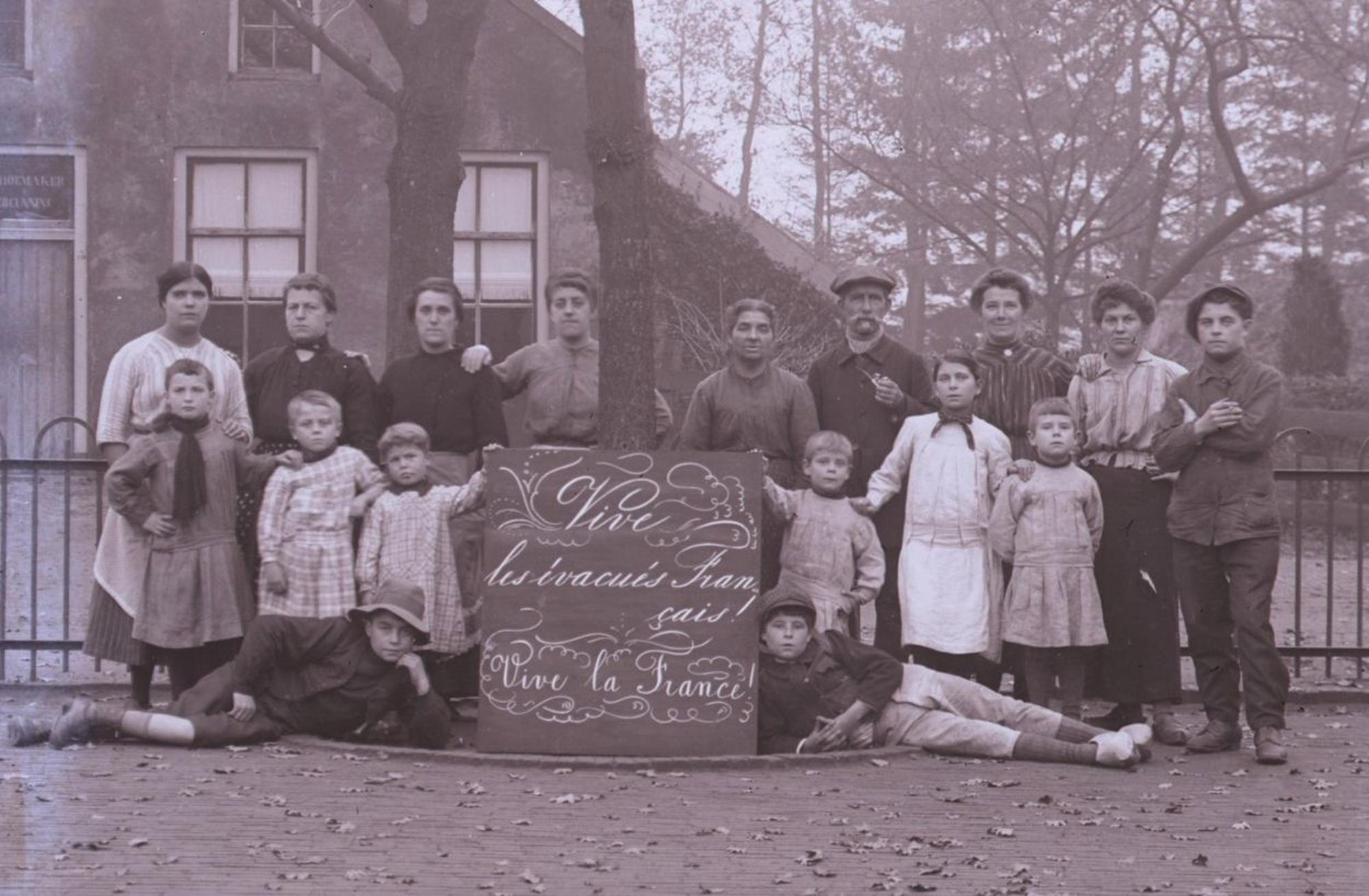 Groepsfoto van kinderen en volwassenen uit het begin van de 20e eeuw, poserend buiten bij een hekwerk. In het midden houden ze een bord vast met de tekst "Vive les braves Français! Vive la France", ter ondersteuning van Frankrijk tijdens de Eerste Wereldoorlog.