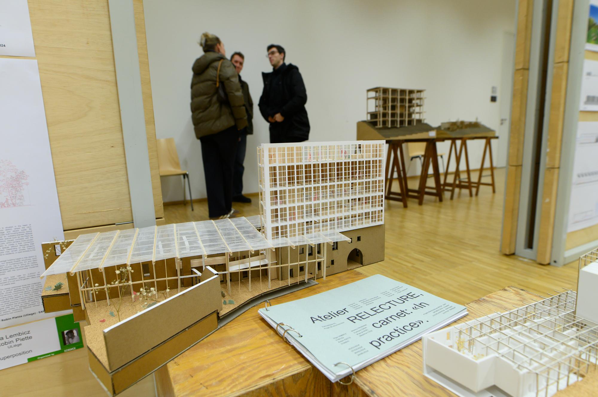 Tentoonstelling van architectuurmodellen op tafels met op de voorgrond een open map van Atelier RELECTURE; op de achtergrond praten drie bezoekers met elkaar.