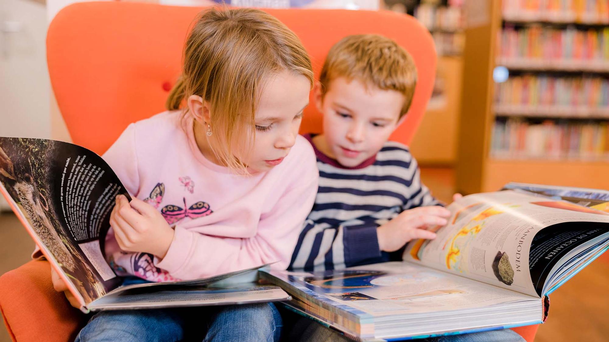 Twee kinderen zitten samen in een oranje stoel en bladeren aandachtig door grote boeken. Op de achtergrond zijn boekenplanken zichtbaar in een bibliotheek.