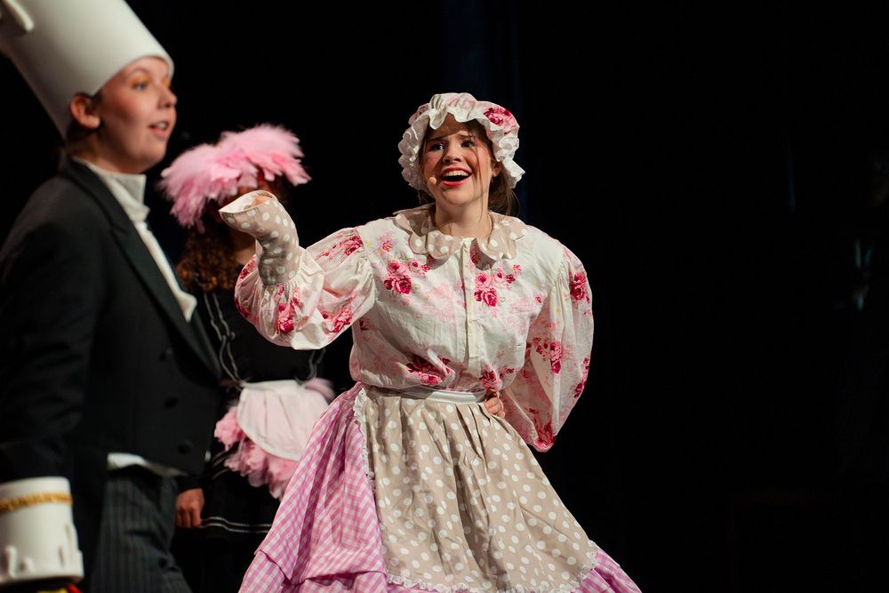Performer in een wit en roze kostuum met bloemenpatroon en muts staat op het podium tijdens de musical Belle en het Beest en maakt een gebaar met de hand. Naast deze persoon staat een performer in een zwart kostuum met een hoog wit hoofddeksel, terwijl op de achtergrond decorstukken en een andere performer met roze veren zichtbaar zijn.
