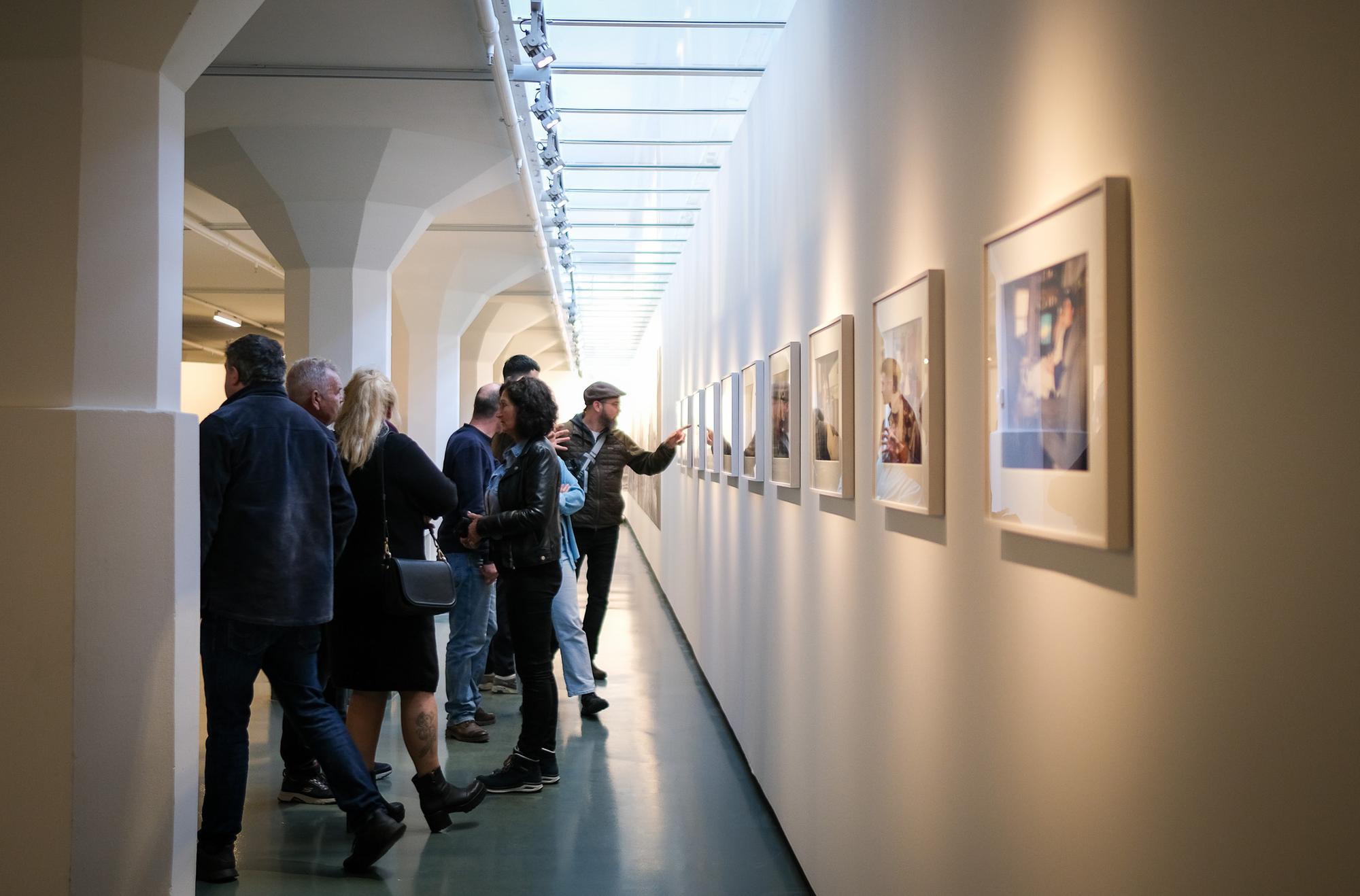 Bezoekers lopen door een gang met fotowerken aan de wand tijdens een tentoonstelling in SCHUNCK, terwijl een man met pet naar een foto wijst.