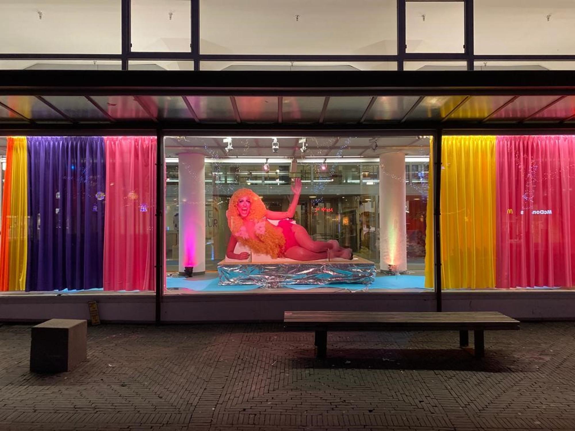 Etalage met kleurrijke gordijnen in geel, roze en paars, waarin een performer met grote gele pruik en roze outfit liggend poseert op een verhoging, met vrolijke verlichting op de achtergrond.