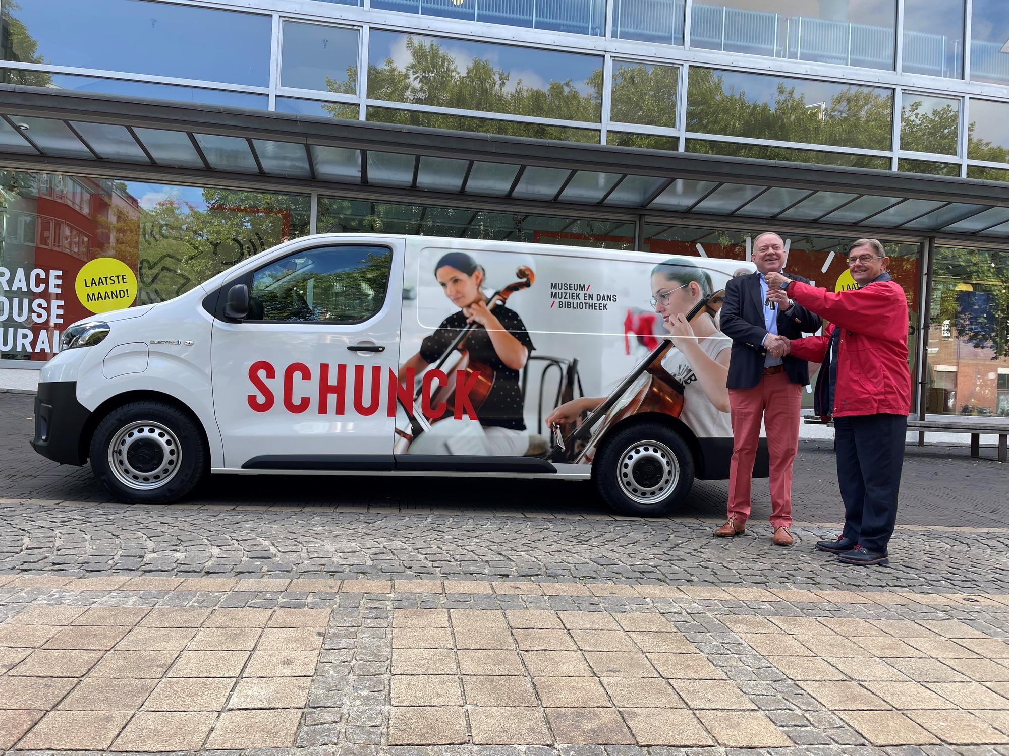 Witte bus met SCHUNCK logo en foto van mensen die cello spelen. Voor de bus staat de directeur met een meneer. 