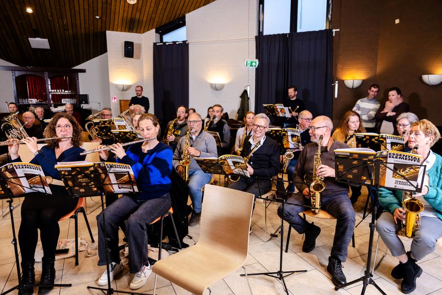 Een orkestensemble repeteert in een zaal met blaasinstrumenten, waaronder saxofoons, trompetten en dwarsfluiten. De muzikanten zitten achter lessenaars met bladmuziek, terwijl enkele toeschouwers achterin de ruimte aanwezig zijn.
