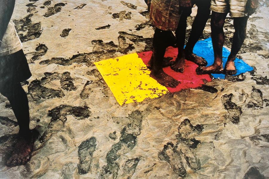Werk van Roy Villevoye met blote voeten van meerdere personen op zand, naast felgekleurde vlakken in geel, rood en blauw.