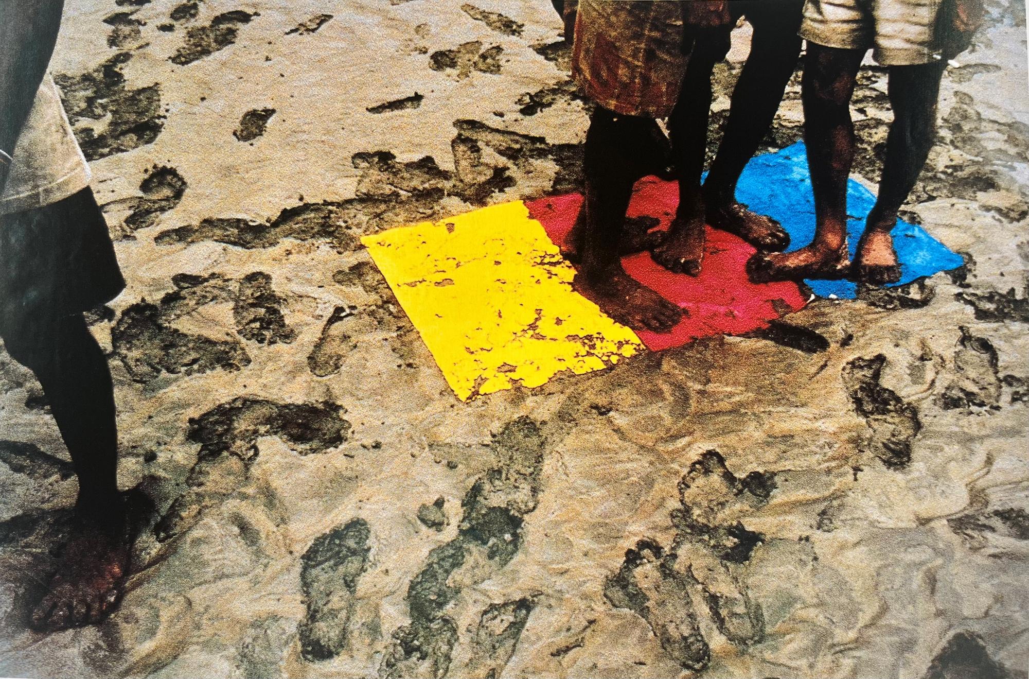 Werk van Roy Villevoye met blote voeten van meerdere personen op zand, naast felgekleurde vlakken in geel, rood en blauw.