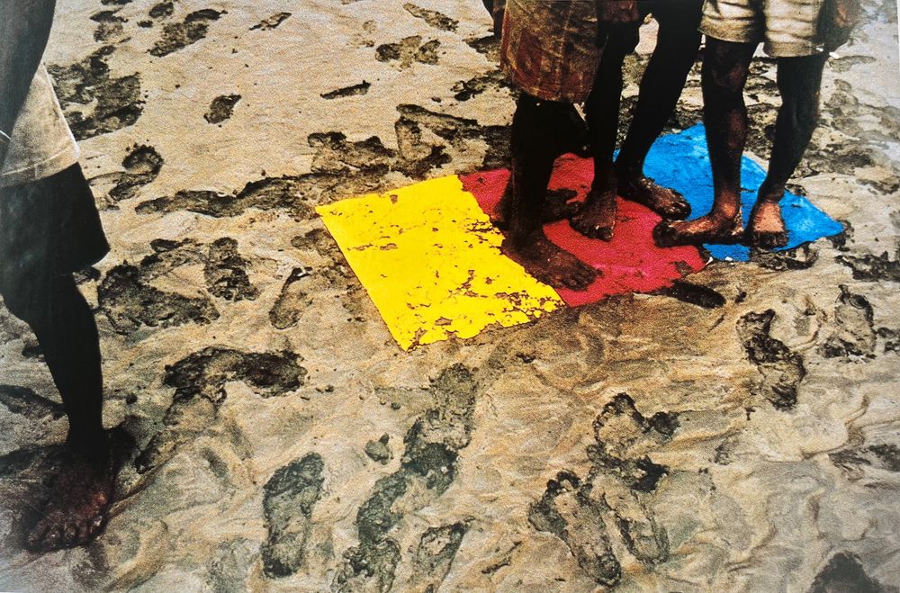 Werk van Roy Villevoye met blote voeten van meerdere personen op zand, naast felgekleurde vlakken in geel, rood en blauw.