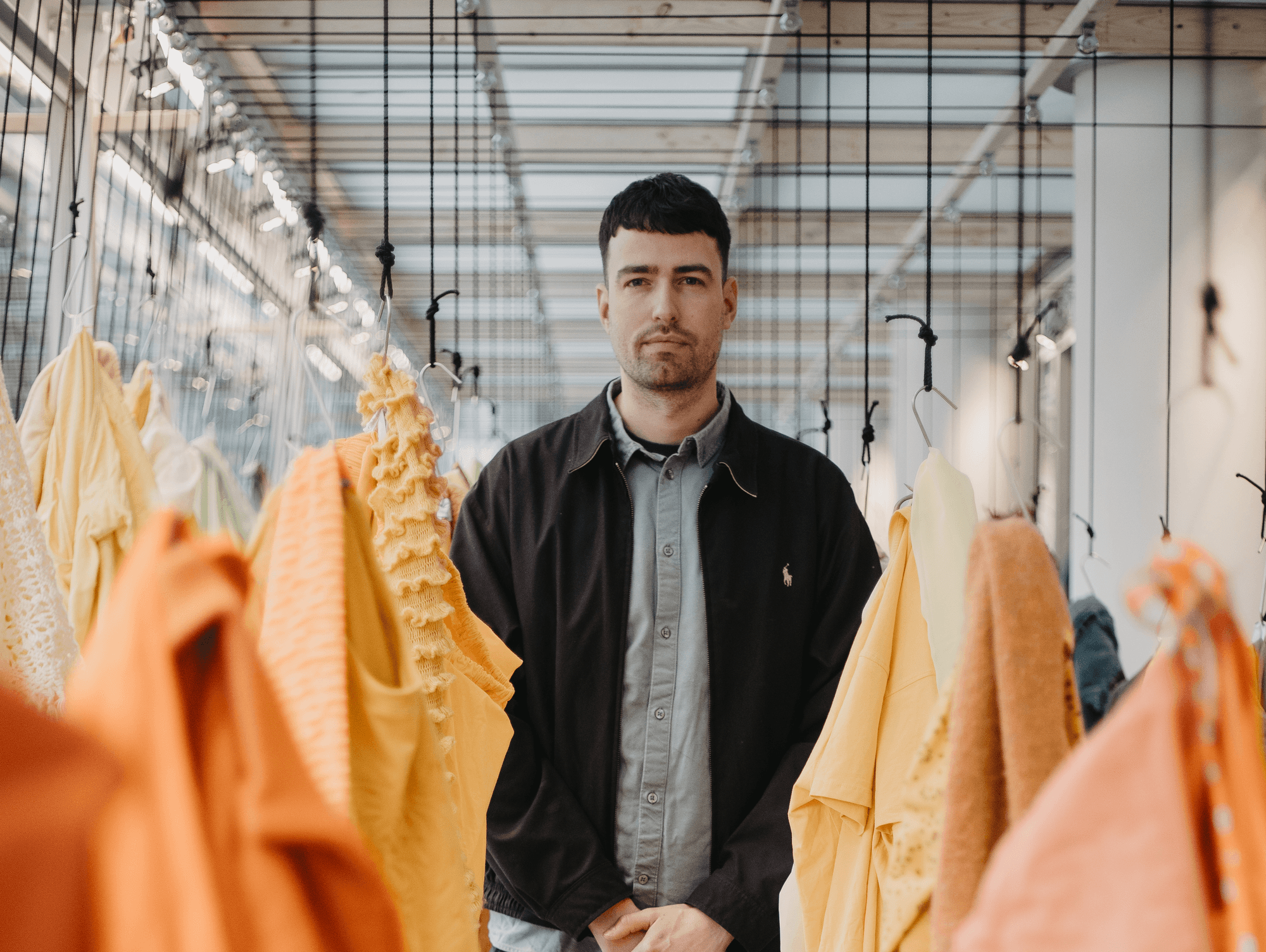 Grondstof: Chaim van Luit SCHUNCK 2023 Man met kort donker haar en een donkere jas staat tussen kleurrijke gele en oranje stoffen die aan hangers in een lichte, moderne ruimte hangen.