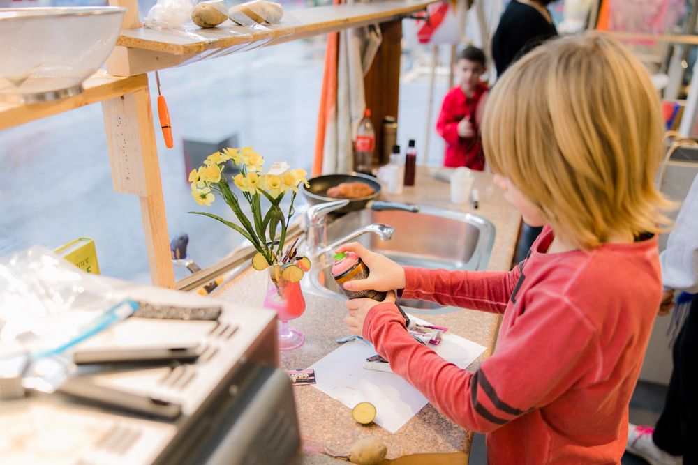 Een kind staat in een keukenachtige workshopruimte en gebruikt een spuitbus terwijl het aan een creatieve opdracht werkt. Op het aanrecht staan bloemen, aardappelen en keukengerei, wat zorgt voor een speelse combinatie van koken, experimenteren en kunst maken.