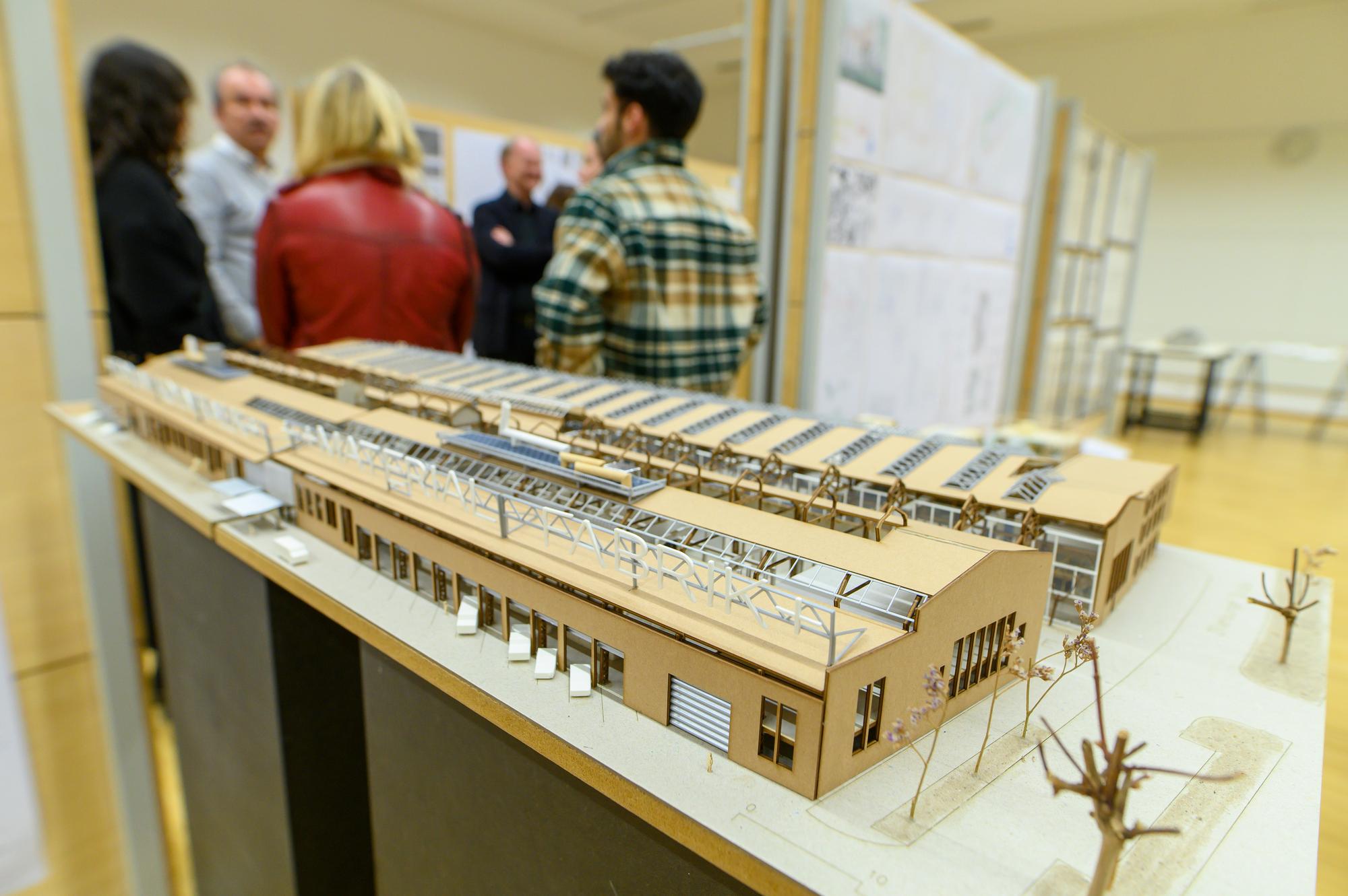 Architectuurmodel van een gebouw tentoongesteld in een expositieruimte, met op de achtergrond een groep mensen in gesprek bij presentatiewanden.
