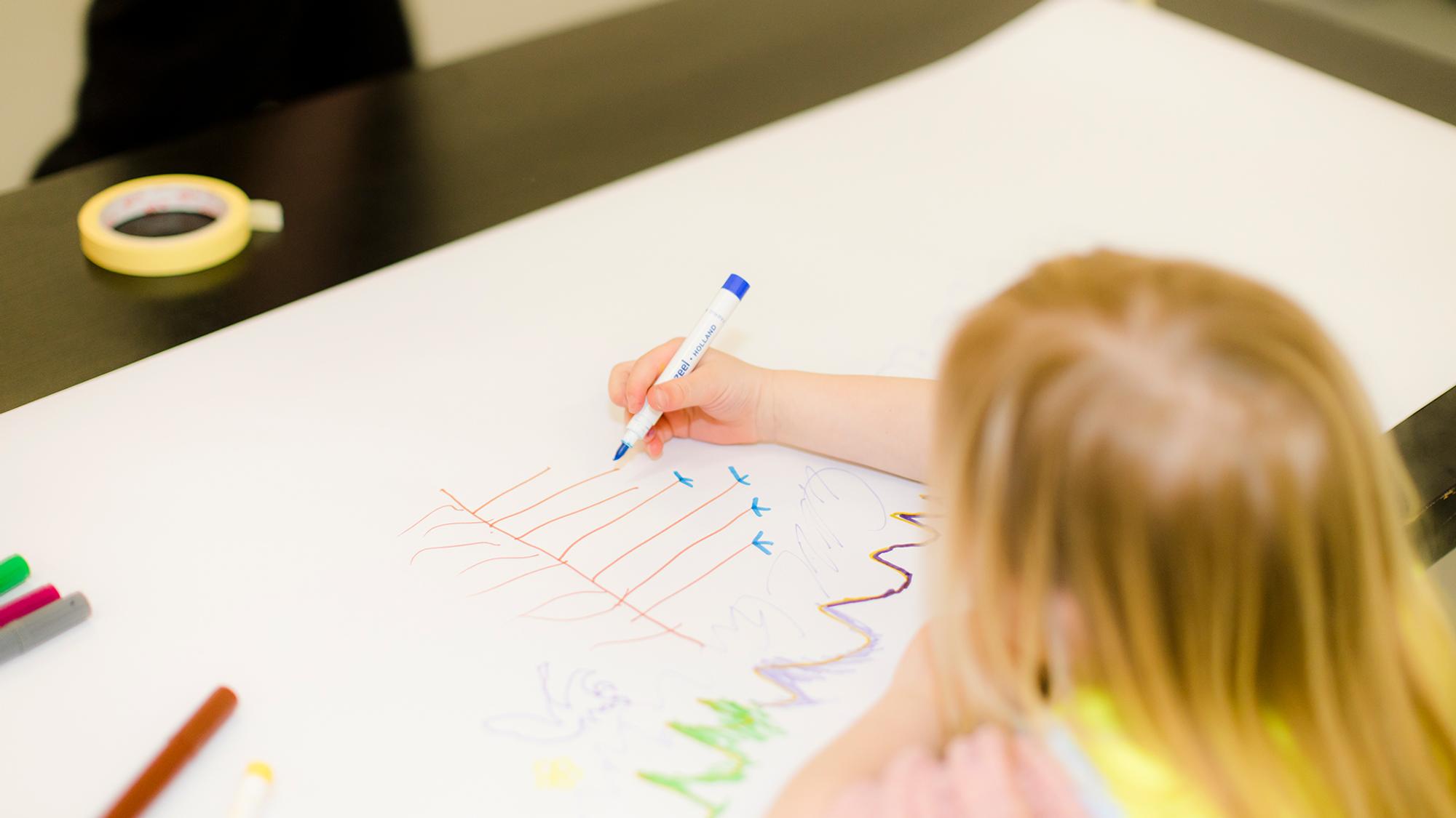 Een kind tekent met stiften op een groot vel papier dat op tafel ligt. Op de tekening zijn gekleurde lijnen, pijltjes en abstracte vormen te zien.