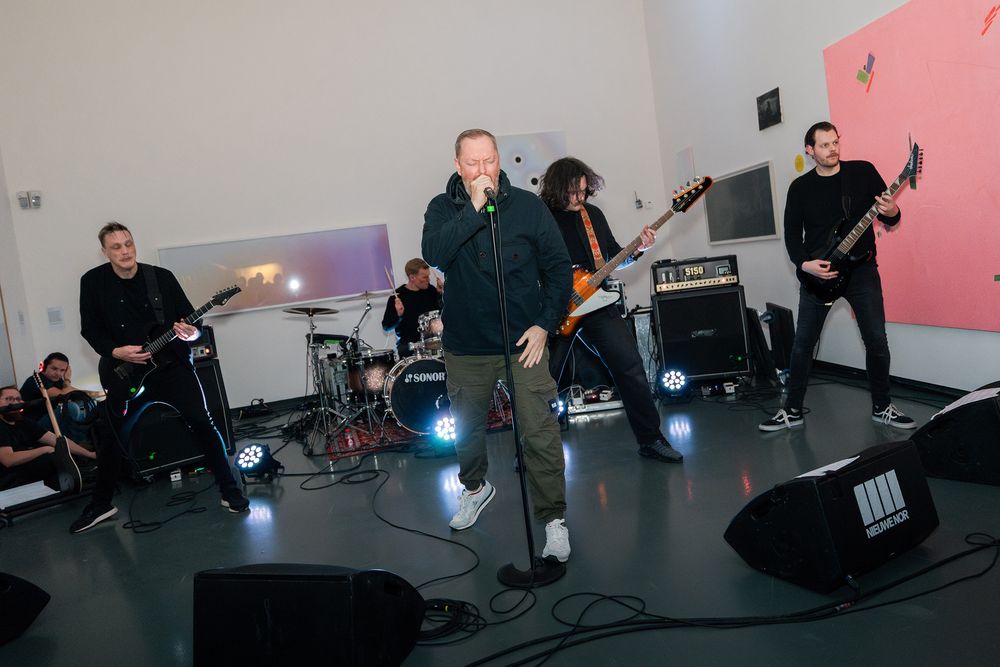Born From Pain (metalband) speelt live in een lichte galerie bij SCHUNCK tijdens Frictions Festival #11; vijf muzikanten staan op het podium met gitaren, drums en microfoon, omringd door versterkers en spotlichten, terwijl kunstwerken aan de muren hangen.
