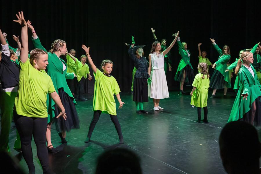 Groep kinderen en jongeren danst op een podium in felle groene en zwarte kostuums, met energieke armbewegingen onder kleurrijk podiumlicht.