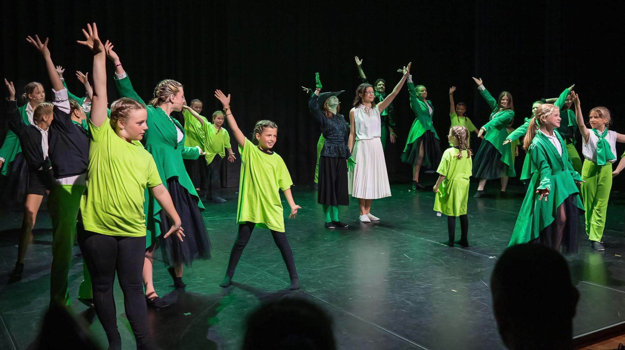 Groep kinderen en jongeren danst op een podium in felle groene en zwarte kostuums, met energieke armbewegingen onder kleurrijk podiumlicht.