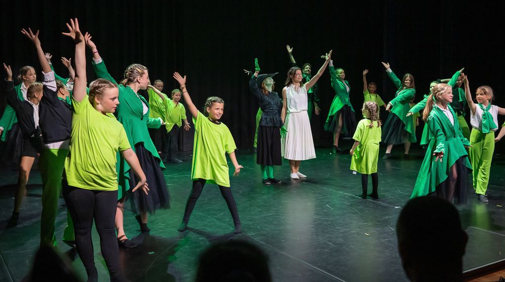 Groep kinderen en jongeren danst op een podium in felle groene en zwarte kostuums, met energieke armbewegingen onder kleurrijk podiumlicht.