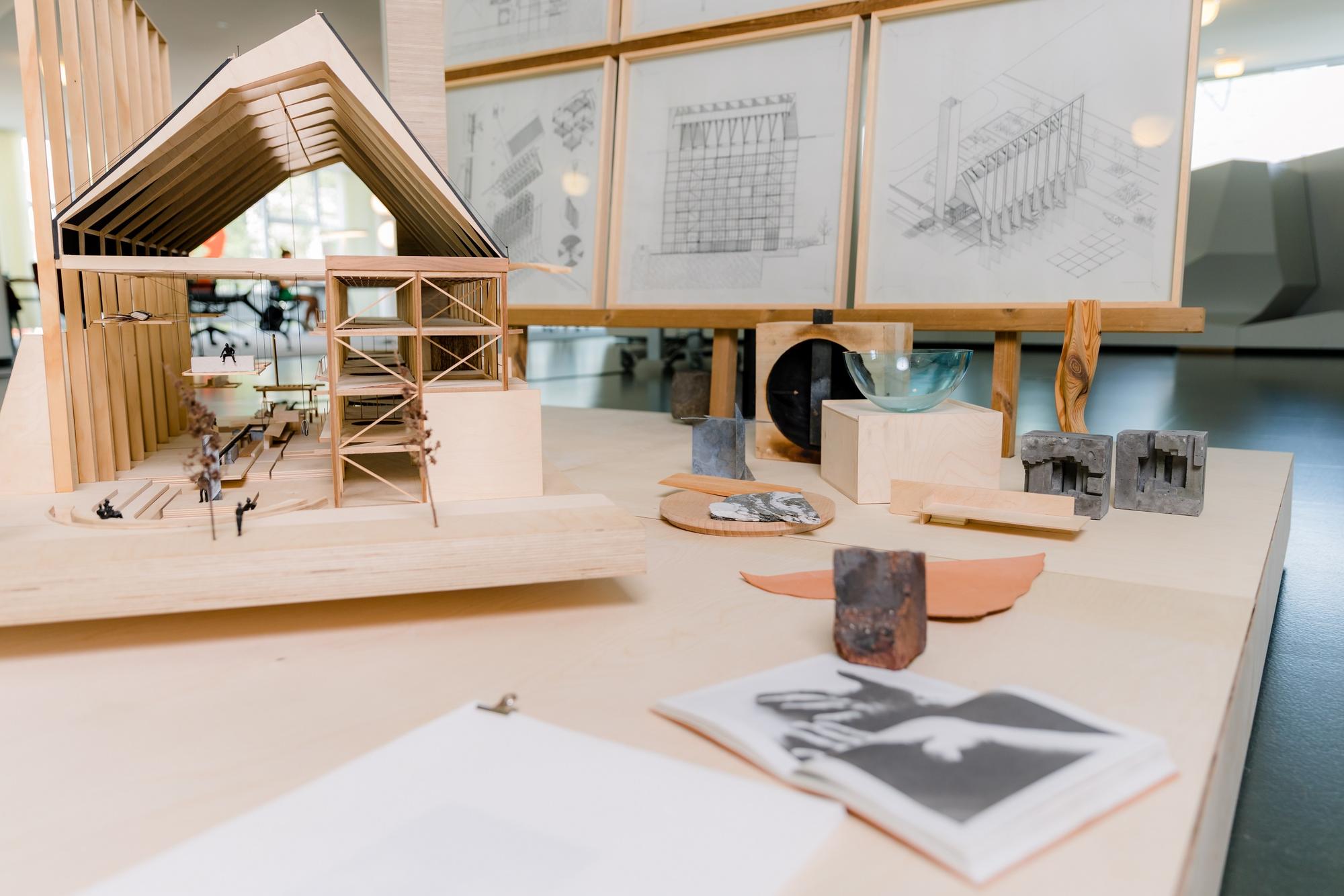 Tafel met gepresenteerde objecten, waaronder een maquette van een gebouw. Op de achtergrond hangen ingelijste tekeningen aan de muur.