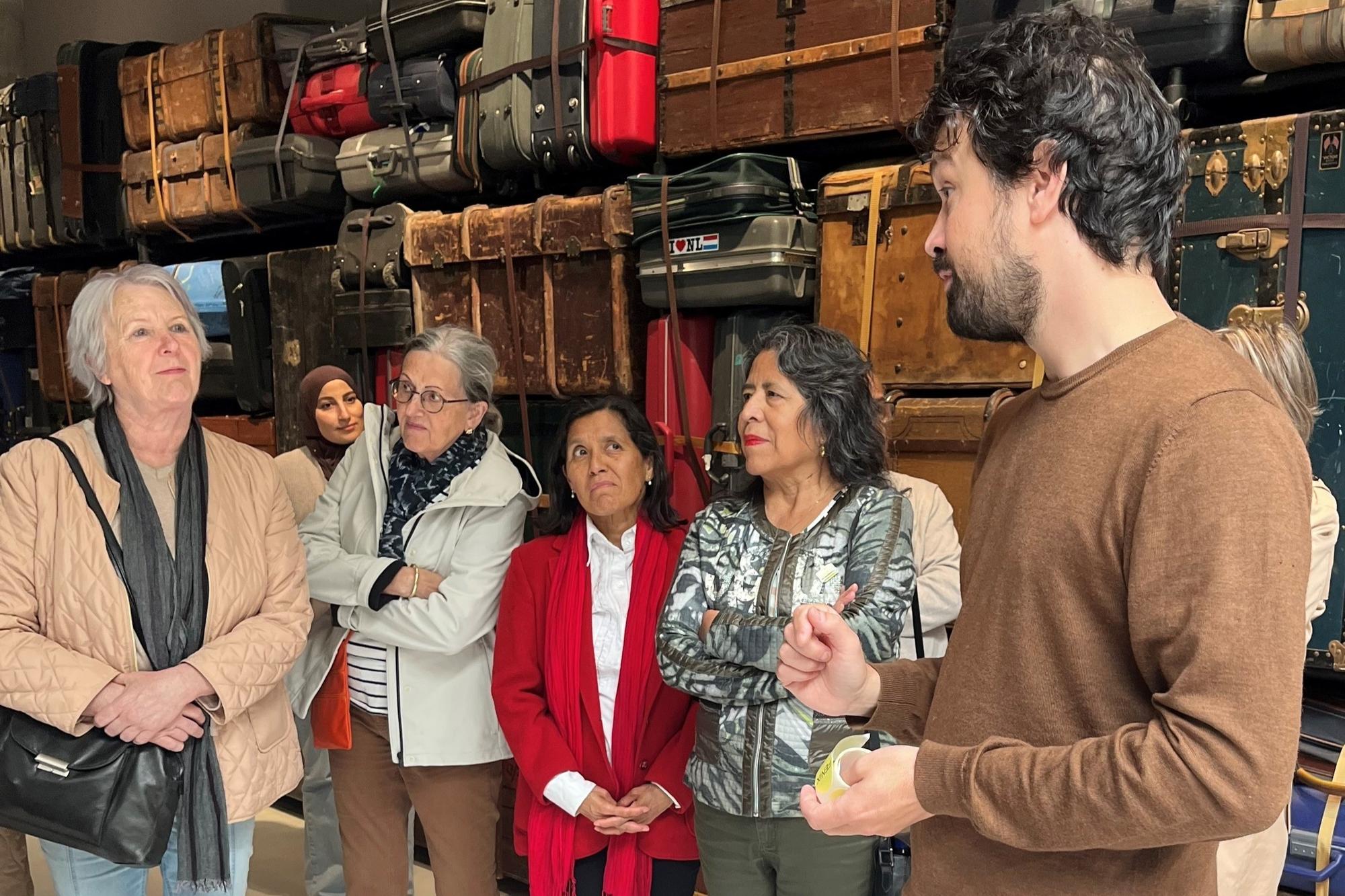 Groep mensen luistert naar uitleg in ruimte met wand vol vintage koffers en reistassen.